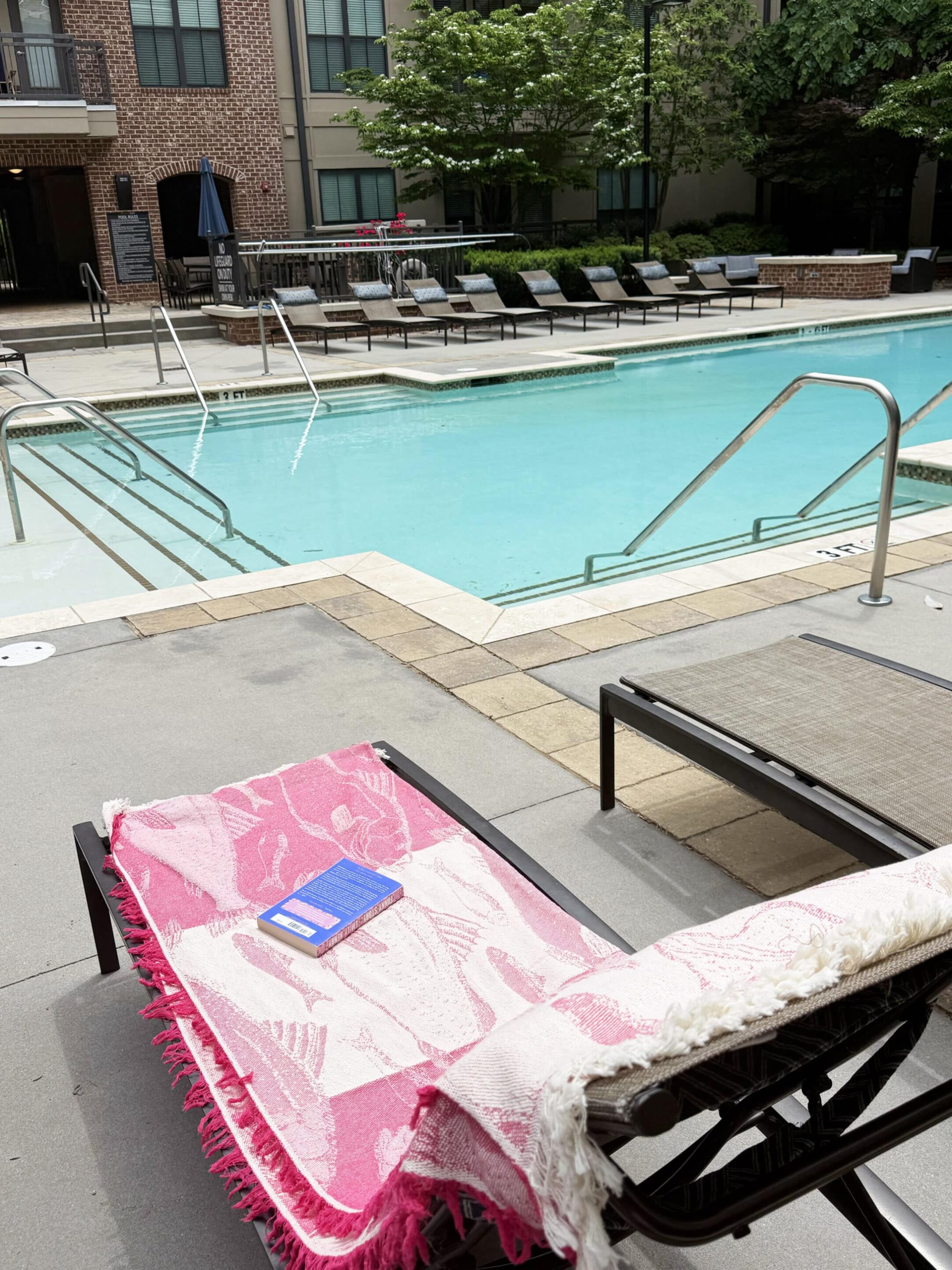 Sandless Blanket 3 Poolside lounge chairs with pink patterned towels and a book resting on one chair by a turquoise pool.
