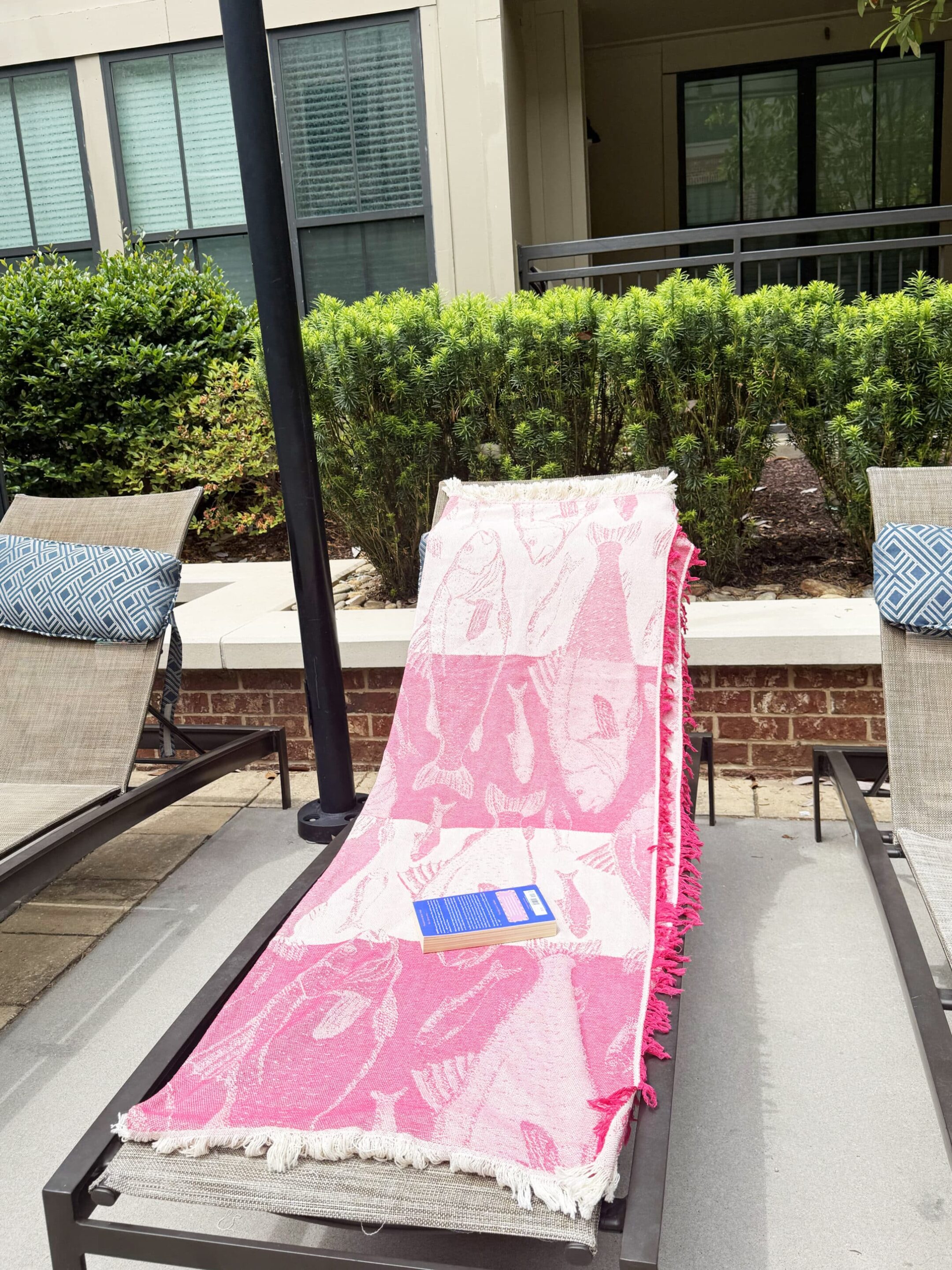 Sandless Blanket 4 Three outdoor lounge chairs with a pink towel on the center chair, a blue book resting on it, and matching blue pillows, backed by shrubs and windows.