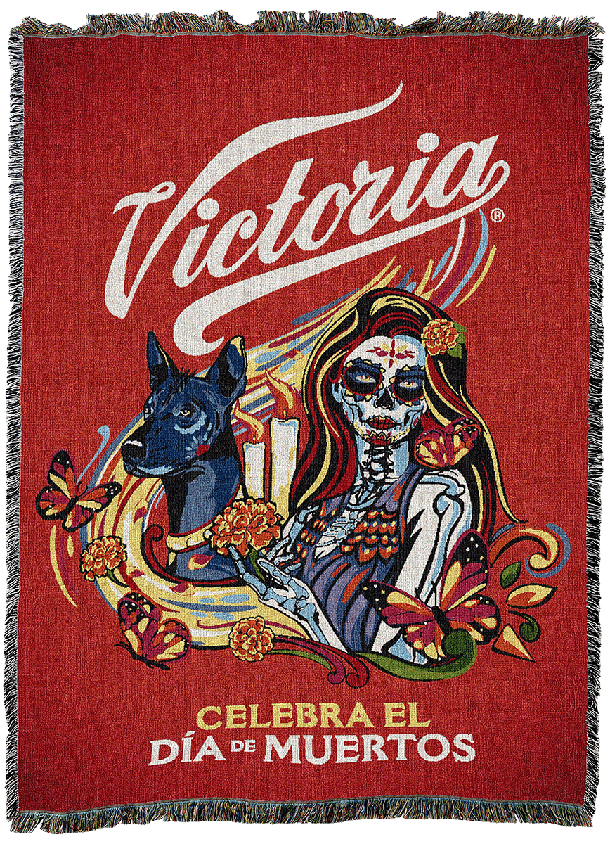 Victoria Tapestry Proof Woven blanket featuring the Victoria beer logo, a person with calavera makeup, a Xoloitzcuintli dog, and marigold flowers with the text CELEBRA EL DÍA DE MUERTOS.